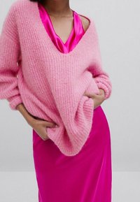 Różowy sweter z dzianiny o luźnym kroju i szerokim dekolcie w serek, nałożony na błyszczącą satynową sukienkę fuksjową. Miękka tekstura i wyraziste kolory.