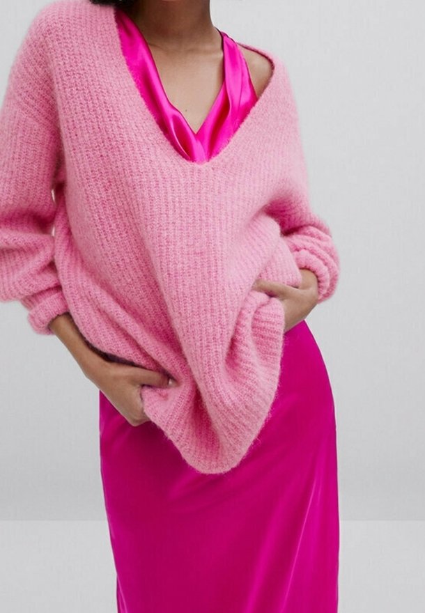 Różowy sweter z dzianiny o luźnym kroju i szerokim dekolcie w serek, nałożony na błyszczącą satynową sukienkę fuksjową. Miękka tekstura i wyraziste kolory.