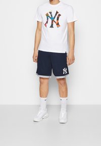 T-shirt blanc avec logo NY coloré, assorti à un short bleu marine avec des bandes blanches et un logo. Des baskets blanches et des chaussettes mi-mollet complètent la tenue.