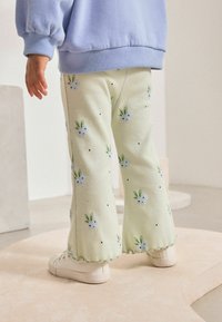 Pantalons en velours côtelé vert clair avec un motif floral, comportant des fleurs bleues et des feuilles vertes. Fini avec un ourlet ondulé et associé à des baskets blanches.