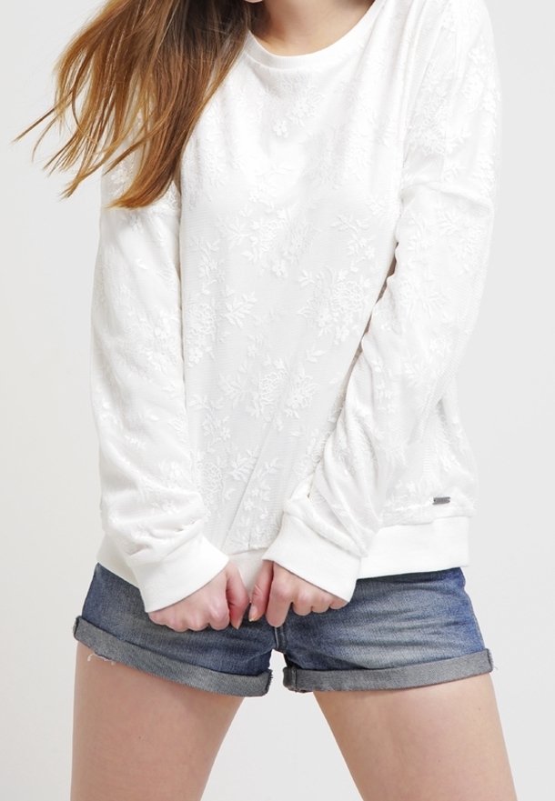 Femme en haut blanc à manches longues avec texture florale et short en jean retroussé, debout, les mains jointes devant elle.