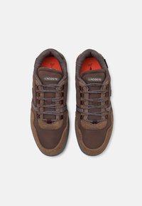 Paire de baskets Lacoste marron en tissu texturé avec des détails en daim, lacets à motifs et semelles intérieures orange vif.