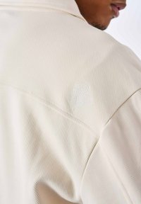 Beige, tekstureret stof med et subtilt ribbet mønster. Har et lille, hvidt broderet logo på skulderen. Skræddersyet pasform og kravedesign.