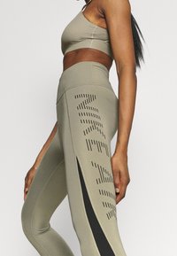 Olivově zelená sportovní podprsenka a legíny s vertikálními perforacemi a černým akcentem na boku, vyrobené z elastického materiálu, který se přizpůsobuje postavě.