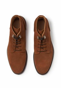 Botas de ante marrón hasta el tobillo con punta redonda, cinco ojales, cordones negros y un forro de cuero contrastante. Suela plana de goma con un acabado texturizado.