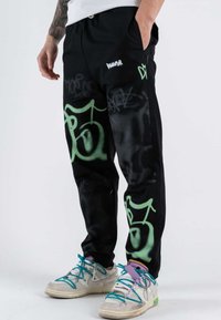 Pantaloni da jogging neri con grafiche verdi simili a graffiti e un logo bianco del marchio. Realizzati in tessuto morbido, con vita elastica e tasche.