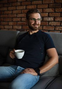 Mand sidder på en grå sofa, iført en marineblå skjorte med knapper og lyseblå jeans, holder en hvid kaffekop op mod en mur af mursten.
