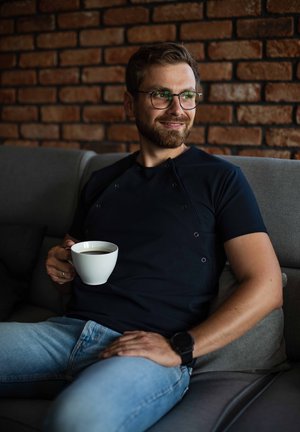 Man zit op een grijze bank, draagt een marineblauw shirt met knopen en lichtblauwe jeans, en houdt een witte koffiekop vast tegen een bakstenen muur.