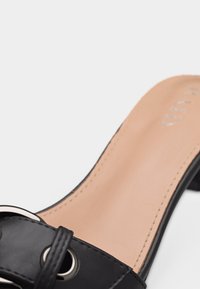 Sandale à glissière en cuir noir avec détail boucle argentée et semelle intérieure beige avec couture visible et talon bas.