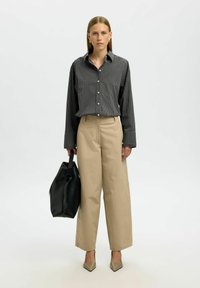 Gestreepte zwart-witte overhemd, beige broek met wijde pijpen, puntige beige schoenen en een zwarte schoudertas met een ronde handgreep.