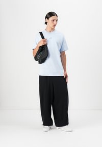 Gaiši zilas kokvilnas t-krekls ar mazu logo, apvienots ar brīviem melniem biksēm un melnu pleca somu, valkāts ar baltām kurpēm.