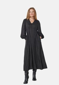 Čierna maxi šaty s dlhými, manžetovými rukávmi a výstrihom do V. Má textúrovanú tkaninu a dve bočné vrecká, doplnené čiernymi čižmami po kolená.