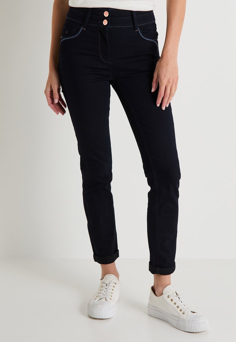 Jeans skinny de mezclilla oscura con dos botones frontales, costuras en contraste y puños enrollados, combinados con zapatillas blancas.