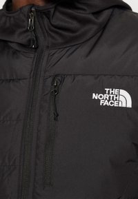 Sort jakke fra The North Face med lynlås foran, tekstureret stof, broderet logo på brystet og hætte-design.