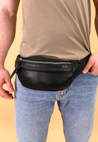 Sort læder bæltetaske med en glat tekstur, lynlåslukning og en rund form, båret om livet over blå denim jeans.
