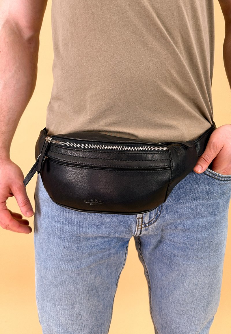 Sort læder bæltetaske med en glat tekstur, lynlåslukning og en rund form, båret om livet over blå denim jeans.