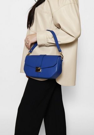 Donna che indossa una giacca beige e una gonna nera, che tiene una borsa in pelle blu con chiusura dorata e tracolla.