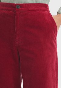 Red corduroy pants featuring vertical ribs, a button closure, and side pockets, displayed against a neutral background.