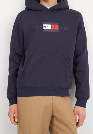 Personne portant un sweat à capuche Tommy Hilfiger bleu marine avec une poche avant et un pantalon beige sur un fond uni.