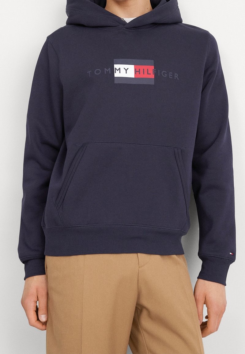 Personne portant un sweat à capuche Tommy Hilfiger bleu marine avec une poche avant et un pantalon beige sur un fond uni.
