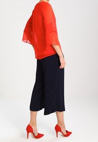 Blouse rouge transparente avec des manches évasées, associée à un pantalon large noir et des talons hauts rouges. Tissu lisse avec une coupe décontractée. Détail de bouton discret dans le dos.