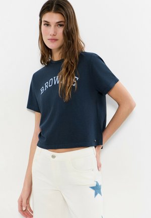 Jeune femme aux longs cheveux bruns portant un t-shirt court bleu marine avec l'inscription "BROWNIE" et un pantalon blanc orné d'un patch étoile bleu.