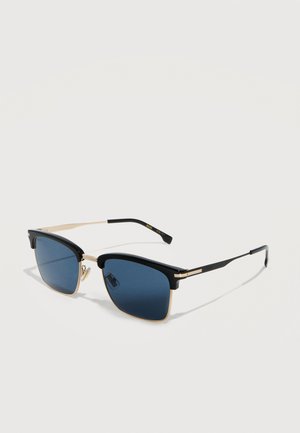 Lunettes de soleil à monture demi-cerclée noire et dorée avec des verres rectangulaires teintés en bleu et des branches fines noires reposant sur une surface blanche.