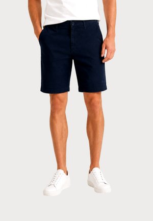 Uomo che indossa pantaloncini blu navy, scarpe da ginnastica bianche e una camicia bianca, con le mani rilassate lungo i fianchi su uno sfondo uniforme.