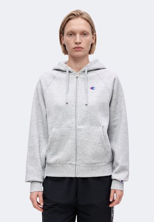 Jong persoon met kort blond haar, draagt een lichtgrijze zip-up hoodie met Champion-logos en zwarte broek, staand tegen een effen achtergrond.