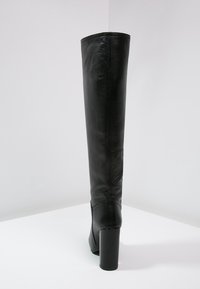 Schwarze Lederstiefel bis zum Knie mit Blockabsatz, aufrecht auf einer weißen Fläche vor einem schlichten hellgrauen Hintergrund platziert.