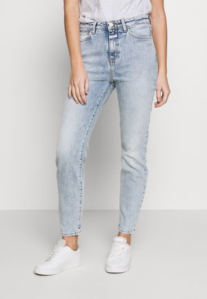 Persoon draagt lichtblauwe jeans met hoge taille, witte sneakers en een in de broek gestoken wit T-shirt, staand tegen een effen achtergrond.