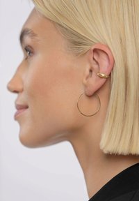 Moteris nešioja auksinį ploną apvalų lankelio formos auskarą ir mažą apvalų ausies segtuką. Lankelis yra plonas ir apvalus; segtukas yra lygus ir apvalus.