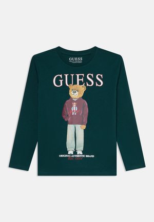 Mørkegrøn langærmet skjorte med en bjørn iført en mørkerød hættetrøje og lyse bukser, tekst "GUESS" ovenover og "Original Authentic Brand Est. 1981" nedenfor.