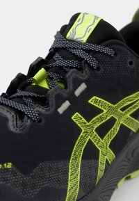 Black athletic shoe with a textured finish. Features yellow accents, patterned laces, and a distinctive logo on the side. Rubber sole.