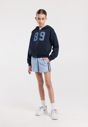 Junges Mädchen trägt einen marineblauen Kapuzenpullover mit Reißverschluss und der Nummer 89, hellblaue Shorts, weiße Socken und weiße Turnschuhe, steht vor einem einfarbigen Hintergrund.