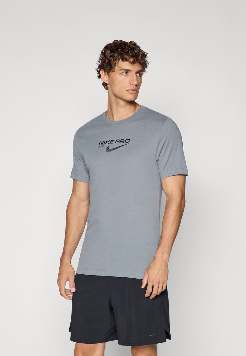 Grå Nike Pro t-skjorte med rund hals, korte ermer og en svart logo trykt på brystet. Stoffet ser glatt og tettsittende ut.
