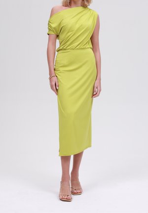 Kobieta w limonkowej, jedno-ramiennej sukience midi z marszczonymi detalami, złotymi bransoletkami i beżowych sandałkach na obcasie stojąca na jednolitym tle.