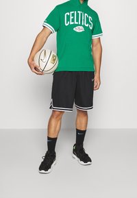 Persona che indossa un hoodie verde dei Celtics, pantaloni corti da basket neri, calze nere Nike e scarpe, mentre tiene un pallone da basket Nike bianco.