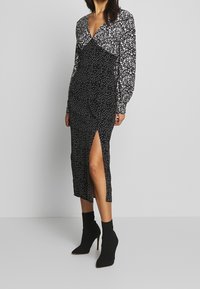 Larga vestido negro con estampado floral blanco, escote en V profundo, mangas largas abullonadas y abertura lateral. La modelo lo combinó con botas negras hasta el tobillo.