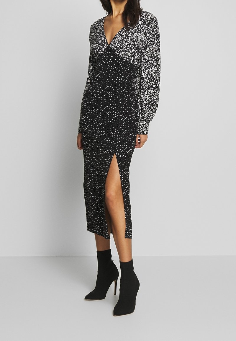 Larga vestido negro con estampado floral blanco, escote en V profundo, mangas largas abullonadas y abertura lateral. La modelo lo combinó con botas negras hasta el tobillo.