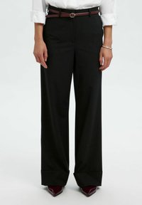 Brede zwart broek met een hoge taille, een stevige textuur en een opgerolde zoom. Gecombineerd met een contrasterende bruine riem en puntige schoenen.