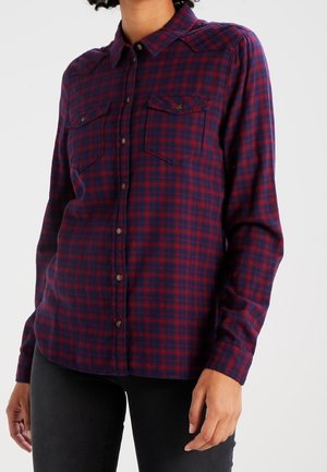 Person trägt ein langärmeliges, rot-navy kariertes Hemd mit Knöpfen und zwei Brusttaschen, kombiniert mit einer schwarzen Hose.