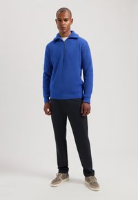 Pulover tricotat albastru cu guler înalt și un fermoar central. Asortat cu pantaloni negri și adidași gri, având o suprafață texturată.