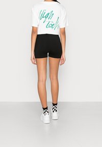 Hvid cropped t-shirt med grøn tekst "High" på ryggen, parret med sorte shorts og ternede sort-hvide sokker, båret med hvide sneakers.