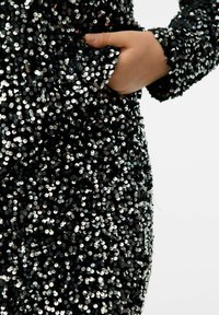 Czarny ubranie z cekinami o błyszczącej fakturze, z boczną kieszenią i długimi rękawami. Cekiny są okrągłe i odbijają światło.