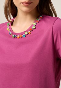T-shirt fuchsia avec un col rond, arborant un ensemble décoratif de perles en plastique colorées et de paillettes le long du col.