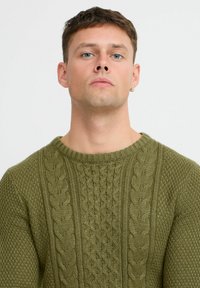 Pulover tricotat de culoare verde măslin, cu un model texturat ce include fire torsadate și un guler rotund. Mâneci lungi, grosime medie și detalii țesute.