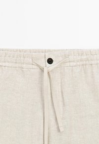 Pantalon en tissu beige avec une taille élastique, un cordon central noué en nœud et un bouton noir au-dessus du cordon.