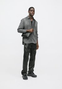 Pantaloni cargo din piele neagră, cămașă gri cu model și butoni, geantă crossbody neagră cu logo și adidași închisi. Texturi netede și croi lejer.