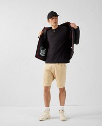 Giacca nera imbottita con fodera rossa, polo nera, pantaloni beige e scarpe da ginnastica chiare. Esibita su uno sfondo neutro.